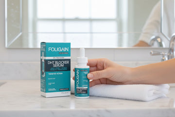 욕실 카운터의 상자와 접힌 수건 옆에서 FOLIGAIN DHT 세럼을 들고 있는 손.